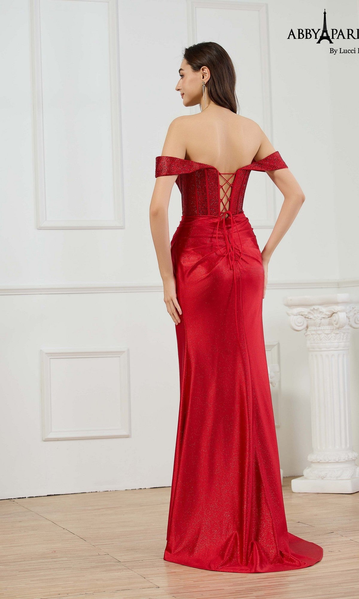 A woman stands indoors wearing the Abby Paris 90382 long prom dress, featuring an off-the-shoulder neckline, fitted corset bodice, lace-up back detail, and a stunning floor-length red gown.